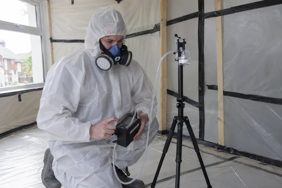 Independent asbestos air-monitoring technician in protective coveralls and respirator adjusting a specialist air-sampling pump connected by tubing to a small filter cassette on a tripod within a controlled empty containment area. Sampler and tubing clearly visible, no dust clouds.
