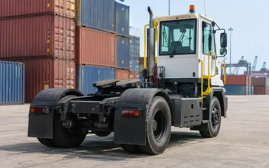 Compact terminal tractor with large rear tyres and an exposed fifth wheel in a container yard.